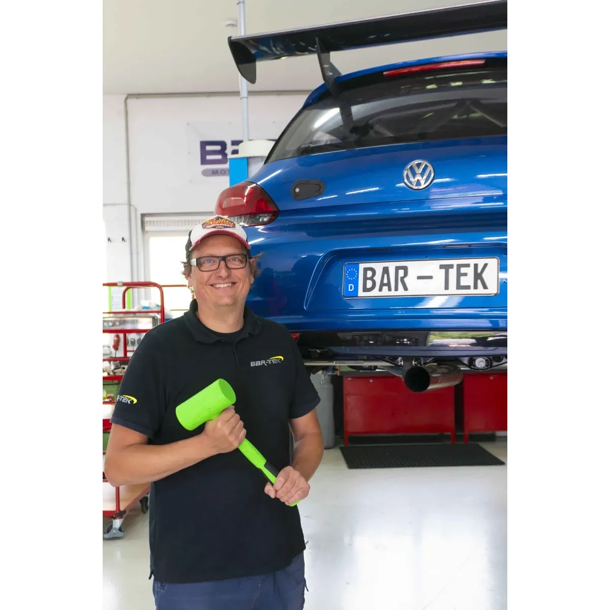 BAR-TEK® Hammer Offer: employee holding a green hammer in front of a blue VW race car on a lift in the workshop