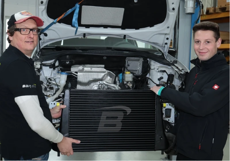 Bartek und Eric halten BAR-TEK® Ladeluftkühler vor Motorblock eines Autos 