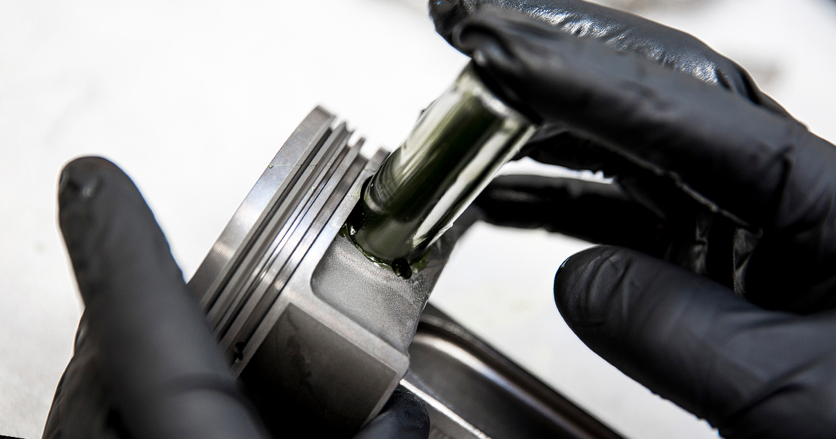 Hände mit schwarzen Handschuhen montieren einen geölten Kolbenbolzen in einen Motor-Kolben – Detailaufnahme eines Motorteils